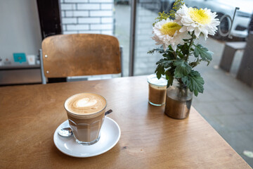 Flat white coffee in clear transparent glass and white plate and flower on the vase, on rough wooden table beside windows blur background of sidewalk.