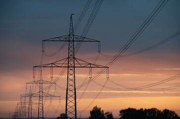 Close-up of large overhead power lines carrying electricity over long distances. The sun rises and shines in the background.