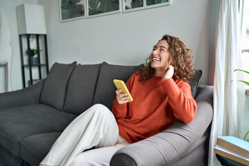 Young happy woman using mobile cell phone sitting on couch at home. Smiling cheerful lady laughing...
