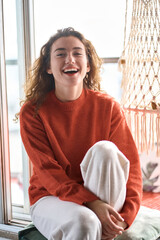 Happy pretty young positive charming woman sitting on windowsill relaxing at home looking at camera. Smiling cheerful lady chilling in apartment, laughing, having fun. Vertical portrait