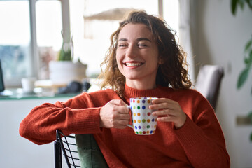 Young smiling pretty woman holding cup drinking warm tea or coffee relaxing dreaming at home. Happy positive lady enjoying hot drink daydreaming with mug in hands in cold cozy winter weekend morning. - Powered by Adobe