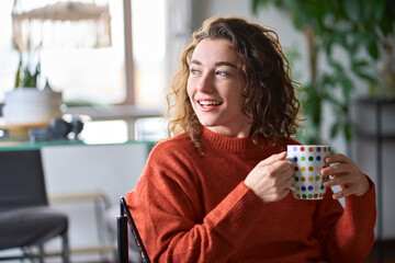 Young smiling pretty woman holding cup drinking warm tea or coffee relaxing dreaming at home. Happy positive calm lady enjoying hot drink with mug in hands daydreaming in cold cozy winter morning.