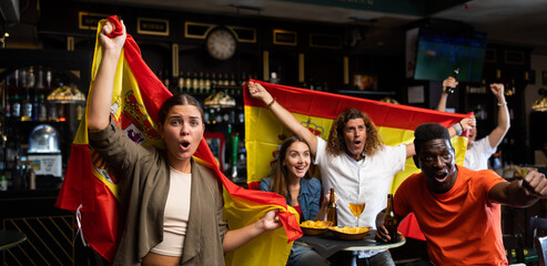 Emotional young girl waving national flag of Spain while watching football match in sports bar,...
