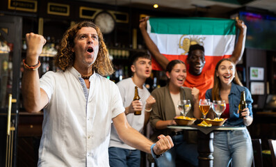 Emotional adult football fan spending time in sports bar, watching game on TV and cheering for favorite team on background of group of cheerful people with national flag of Iran ..