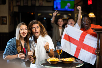 Man and woman, England football team fans, spending time in bar, drinking bear and screaming chants. People with state flag in pub.