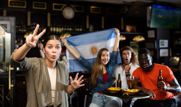 Excited young woman, Argentina football team fan, spending time in bar with friends, making victory sign with fingers. People with state flag in pub.