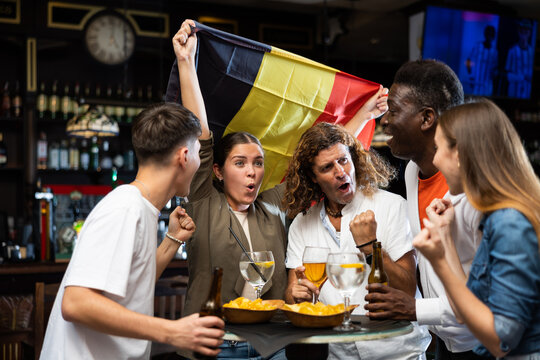 Excited diverse soccer fans with flag of Belgium celebrating victory of team with pint of beer and chips in the pub