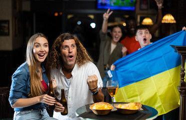 Obraz premium Expressive adult man sitting with cheerful girlfriend at table in sports bar, watching football match and emotionally rooting for favorite team while on background of fans with Ukrainian flag..