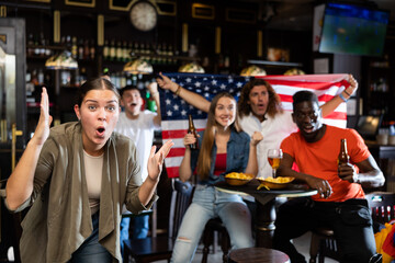 Excited young woman, USA football team fan, gesturing and screaming chants. People with state flag in pub.