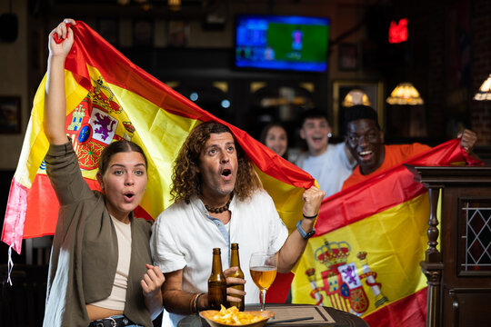 Excited couple watching football match with interest and anticipating goal of favorite team at table in sports bar, holding national flag of Spain