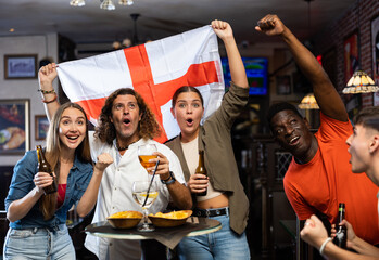 Excited young adult friends, sports fans celebrating victory of favorite football team with beer in...