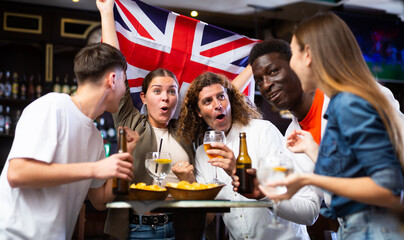 Multiracial United Kingdom sports fans, men and women, supporting their favourite team in bar, raising state flag and screaming chants together.