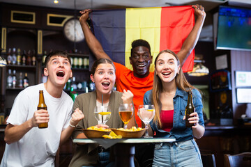 Excited diverse soccer fans with flag of Belgium celebrating victory of team with pint of beer and...