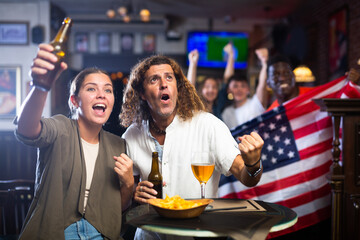 Expressive young adult couple sitting at table in sports bar, drinking beer and emotionally...