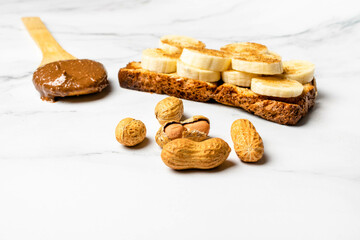 Wooden spoon containing chocolate peanut butter, cocoa and vegan brownie next to a toast with sliced banana and cinnamon on top with peanuts on the side. Mention healthy eating and daily care.
