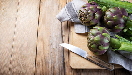 Raw artichokes, cutting board and knife on wooden background, close-up, space for text.