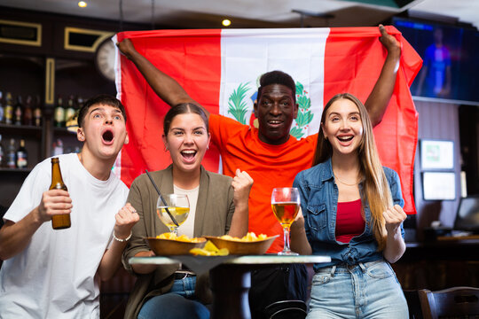 Multiracial Peru sports fans, men and women, supporting their favourite team in bar, raising state flag and screaming chants together.