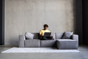 Smiling ethnic indian man sitting at home on sofa using laptop looking at computer working or...