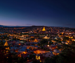 Naklejka premium Summer night view on the city of Tbilisi Tiflis the capital of Georgia with streets of Old Tbilisi in foreground, Mtkvari Kura river, Holy Trinity Cathedral, Peace Bridge, State Palace of Ceremonies