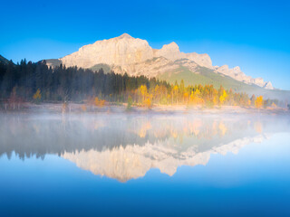 Fototapeta na wymiar Mountain landscape at dawn. Foggy morning. Lake and forest in a mountain valley at dawn. Natural landscape with bright sunshine. Reflections on the surface of the lake. Alberta, Canada.