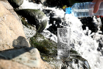 Woman pouring water from plastic bottle into glass near stream outdoors, closeup. Space for text