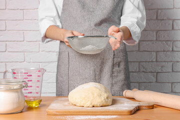 Woman sprinkling flour over dough at table near white brick wall, closeup