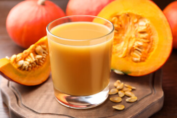 Tasty pumpkin juice in glass, whole and cut pumpkins on wooden table, closeup