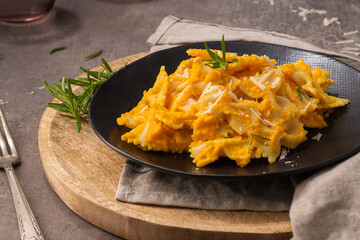 Farfalle pasta with pumpkin sauce and parmesan cheese decorated with rosemary on a black ceramic plate. Creamy cheesy vegan pumpkin pasta. Kitchen counter top.