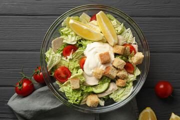 Bowl of delicious salad with Chinese cabbage, lemon, tomatoes and bread croutons on black wooden table, flat lay