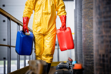 Unrecognizable worker in hazmat protection suit walking by chemicals reservoirs and carrying...