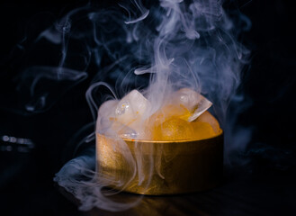Golden jar containing ice cubes or orange ice on a dark background with the cold smoke.




