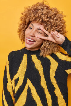Positive good looking European woman with curly hair winks eye smiles broadly makes peace gesture sticks out tongue enjoys life wears comfortable black and yellow jumper poses in studio alone