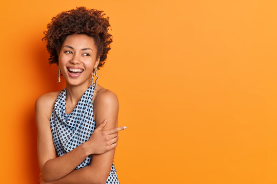 Cheerful curly haired woman keeps arms crossed over body looks gladfully aside smiles broadly wears star shaped earrings and polka dot blouse has healthy clean skin isolated over orange background