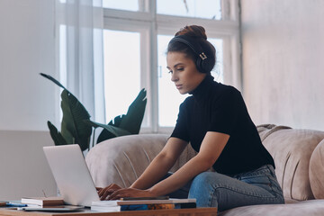 Confident young woman in headphones using laptop while sitting on the couch at home
