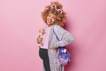 Positive curly haired pregnant woman holds doll carries net bag full of toys dressed in casual clothes enjoys happy expectations poses for article about successful mothers isolated on pink background