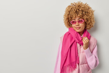 Thoughtful dreamy woman with curly hair looks pensively aside wears stylish sunglasses vest and scarf around neck concentrated aside isolated over grey background blank space for your promotion