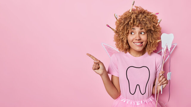 Horizontal shot of tooth fairy poses with magic wand points away on blank space recommends to visit dentist bites lips dressed in costume with wings isolated over pink background. Symbol of childhood