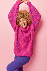 Vertical shot of joyful carefree young woman dressed in knitted loose jumper and trousers keeps arms raised up dances feels very happy isolated over pink background. Positive emotions concept