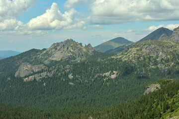 High cliffs of mountain ranges with coniferous forest at the foot under a cloudy summer sky.