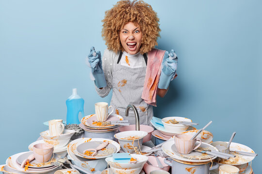 Emotional housewife yells angrily does housework has to wash up surrounded by pile of dirty dishes uses chemical detergent busy with housekeeping isolated over blue background. Dishwashing concept