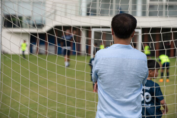 Dad standing and watching his son playing football in a school tournament on a sideline with a...