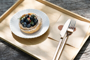 Blueberry tart with knife and fork on wooden tray in natural light.