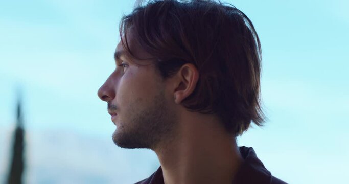 Young man looks around standing against blurry blue sky. Bearded guy searches important things and thinks about life at evening dusk closeup