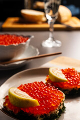 two sandwiches with red fish caviar served with and a slice of lemon, a glass of champagne in the background a porcelain white bowl filled with caviar with a teaspoon and sliced wheat bread