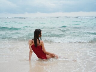 Woman with a beautiful tan tourist in a red swimsuit sitting on the sand on the beach in the ocean in the waves with her back to the camera, cloudy weather storm