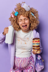 Frustrated unhappy pregnant woman holds bottle of milk and sweet doughnuts has overeating habits during pregnancy has dirty clothes poses against purple background going to give birth to baby