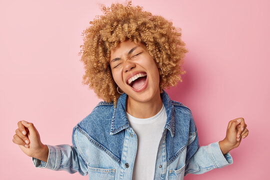 Overjoyed curly haired woman dances carefree keeps arms raised keeps mouth opened exclaims loudly wears stylish denim jacket isolated over pink background has upbeat mood. Positive emotions.