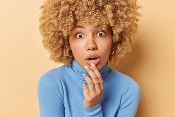 Headshot of amazed curly haired woman keeps mouth opened hand near lips stares with omg expression at camera wears blue turtleneck isolated over beige background cannot believe in shocking news