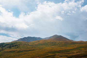 Scenic motley autumn landscape with sunlit high hill top and mountain range under cloudy sky. Fading autumn colors in mountains. Dramatic scenery with sunlight under rainy clouds in changeable weather