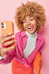 Joyful pretty woman winks eye sticks out tongue takes selfie via smartphone wears white turtleneck and shirt poses with small bag has video conversationn with best friend isolated over pink background